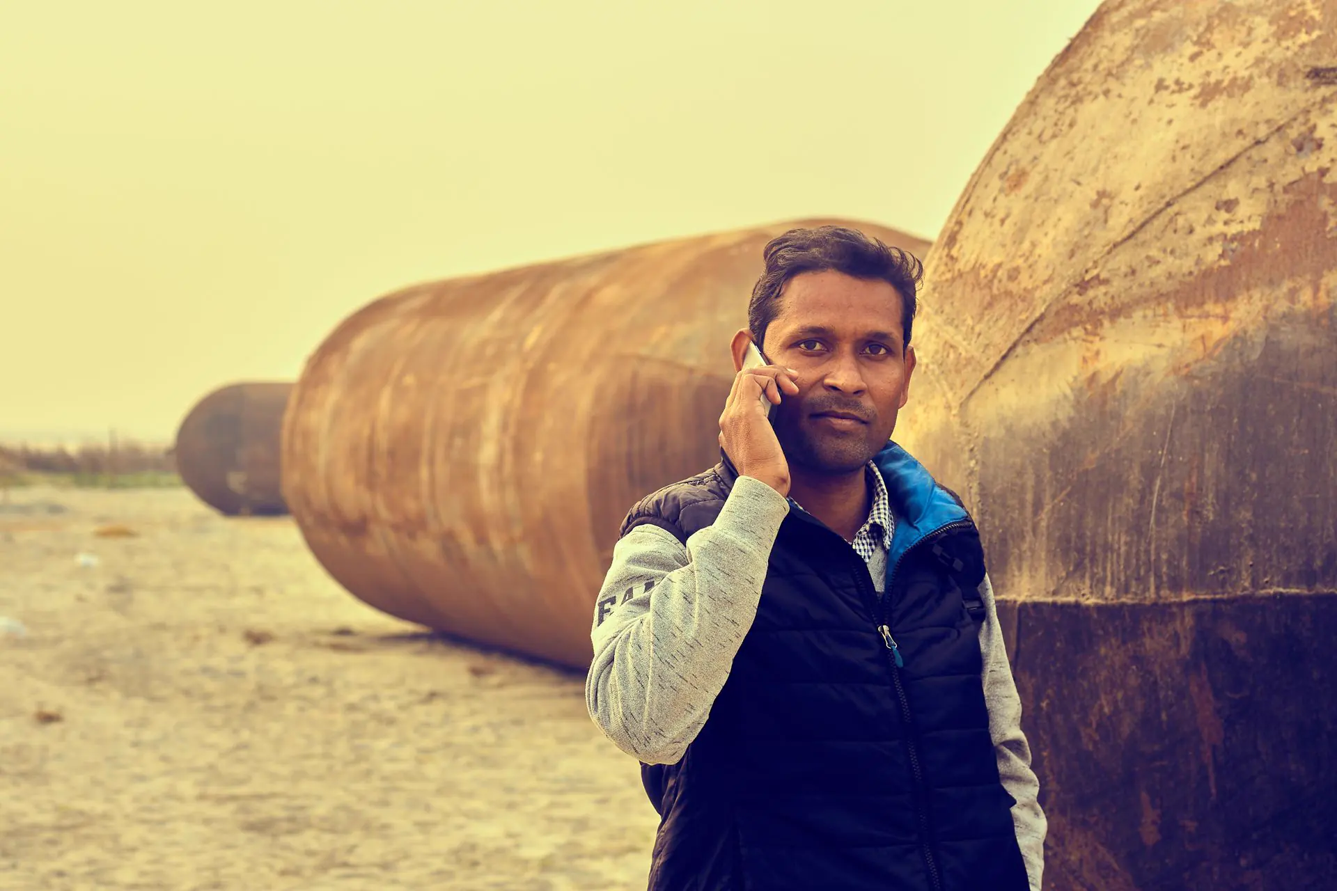 A man in a work jacket taking a phone call beside a row of large rusted industrial pipes
